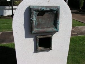 A small, weathered bronze sculpture of a window on a rounded white pedestal.
