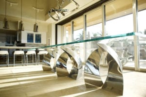 Modern dining table with unique metallic base and glass top in a sunlit room.