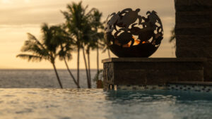 Decorative fire bowl glowing by the pool at sunset with palm trees in the background.