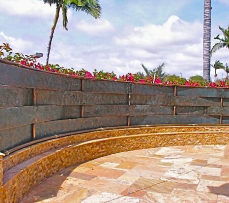 Curved stone wall with tropical plants and a tiled patio under a blue sky.