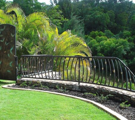 A sunlit garden with green grass, palm trees, and a stone-bordered curved pathway.