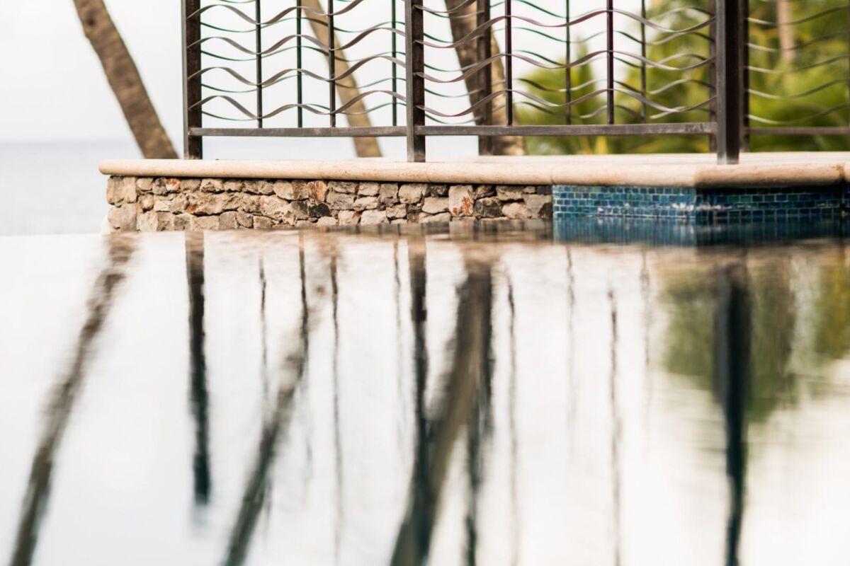 Reflections of palm trees on a calm pool surface near a stone wall.