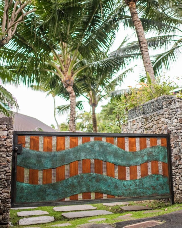 Decorative gate with wavy metal design surrounded by palm trees and stone walls.