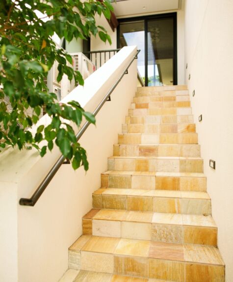 Sunlit stone stairs with a black handrail and greenery nearby.