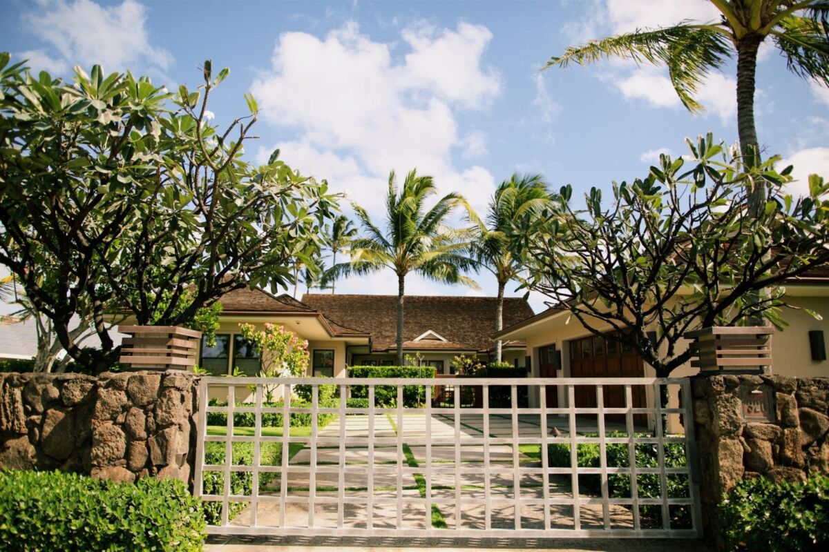 A tropical house with palm trees behind a white gate under a blue sky.