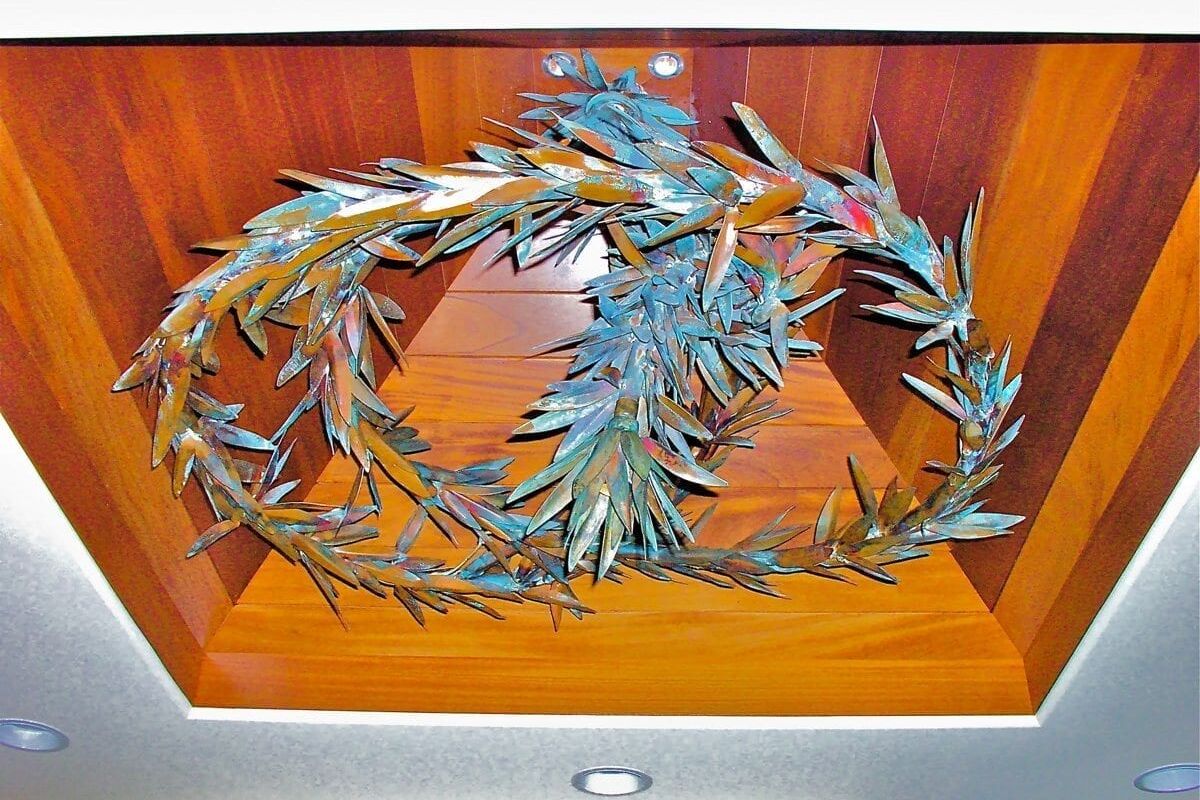 Decorative metal olive branch wreath mounted on a wood panel.