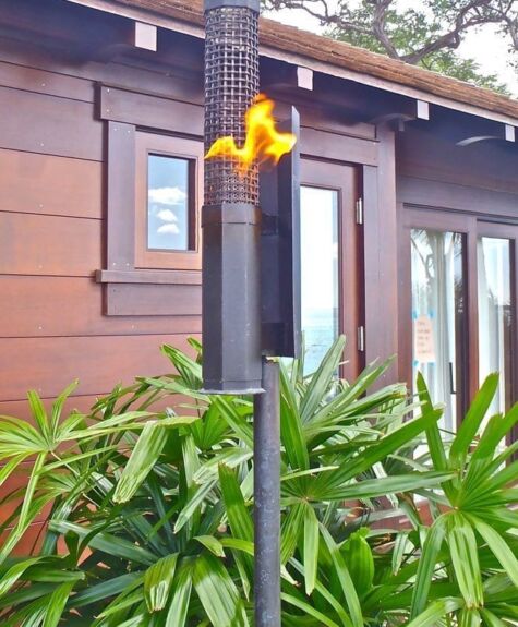 A tiki torch burning outdoors near a wooden house and green plants.