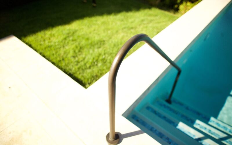 View of a blue swimming pool ladder and green grass in sunlight.