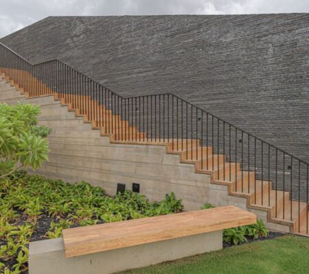 Concrete stairs with a wooden handrail beside a garden bench and greenery.