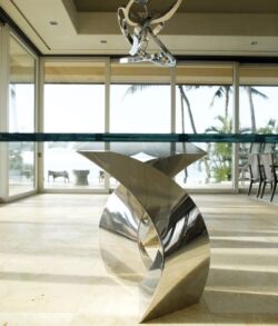 Modern silver whale tail sculpture in a sunlit room with large windows.