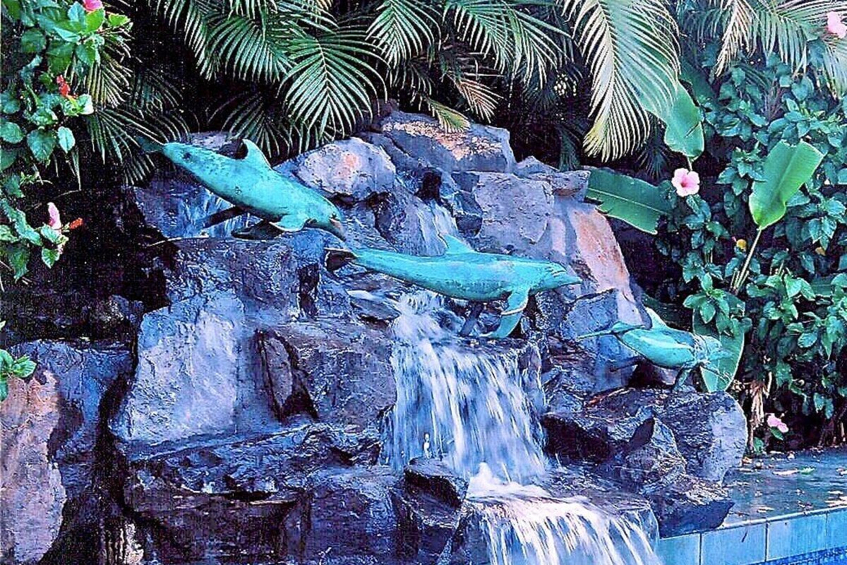 Three green dolphin sculptures leap over a cascading waterfall in a tropical setting.