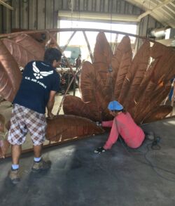 The main entry gate with the texture of the leaves with all natural copper colors Two people assembling a large metal sculpture resembling a leaf indoors.