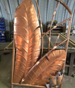 The pedestrian gates taking shape before bamboo leaves and patina Two large, copper-colored metal feathers in a workshop.
