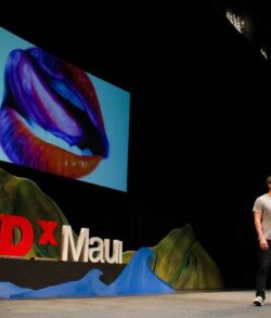 Kamea Hadar guest speaker- The Catalytic Nature of Art Speaker presenting at TEDxMaui with a colorful dinosaur image projected behind.