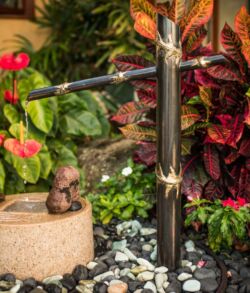 Copper bamboo water feature A small water fountain with a metal spout and a stone basin surrounded by plants.