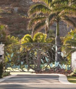 Copper and brass floral and foliage main entrance gate A gated residential entrance with palm trees and lush landscaping.
