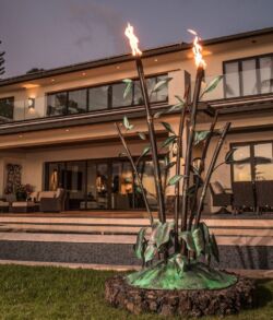 Free standing copper bamboo and philodendron fire feature A tiki torch setup in front of a modern house at dusk.