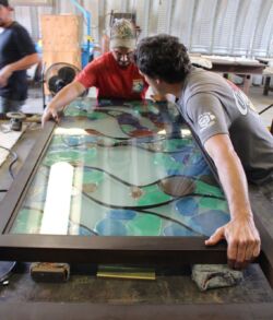 Two people working on a stained glass window frame in a workshop.