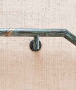 Our classy signature copper and brass bamboo handrail featuring our custom brushed green patina coating Close-up of a metallic handrail mounted on a textured wall.