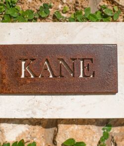 Rusty metal plaque with the name 'KANE' on a stone base.