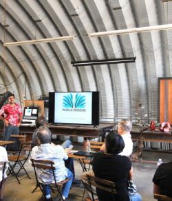 Kaleo Padilla presenting Padilla Designs to attendees People attend a presentation in a rustic, curved-roof room with a digital screen.