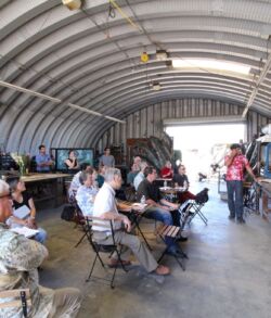 People seated inside a large hangar, listening to a speaker.