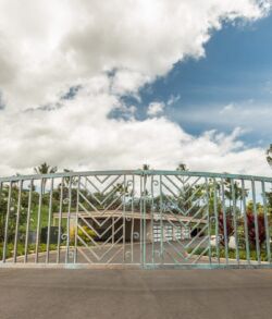Closed metal gate guarding a driveway with greenery and cloudy sky.