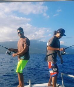 Two men fishing on a boat in calm ocean waters under a clear blue sky.