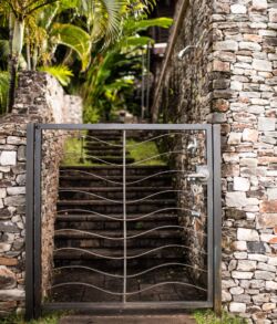 Modern metal gate with stone walls and lush tropical plants behind it.