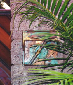 Framed stained glass window partially obscured by green palm leaves on stone wall.