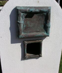A small, weathered bronze sculpture of a window on a rounded white pedestal.
