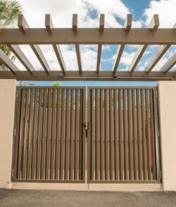 Modern metal gate with horizontal slats under a pergola against a blue sky.