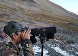 Person in camouflage using binoculars on rocky terrain.