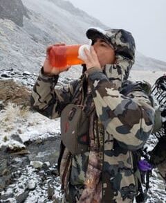 A person in camouflage gear drinking from a bottle outdoors.