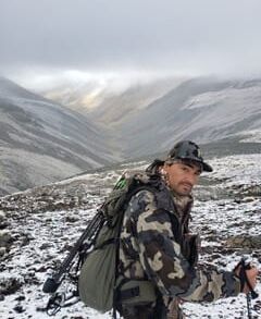 Man in camouflage gear hiking through snowy mountainous terrain.