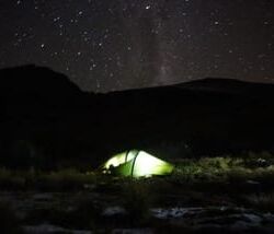 A glowing tent under a starry night sky in a mountainous area.