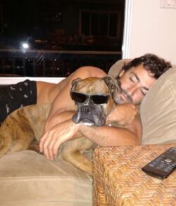 Man cuddling a dog wearing sunglasses on a couch.