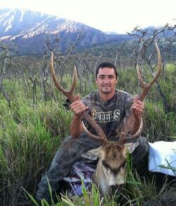 Man posing with a large deer in a scenic outdoor setting.