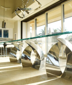 Polished Stainless steel table base Modern dining table with unique metallic base and glass top in a sunlit room.